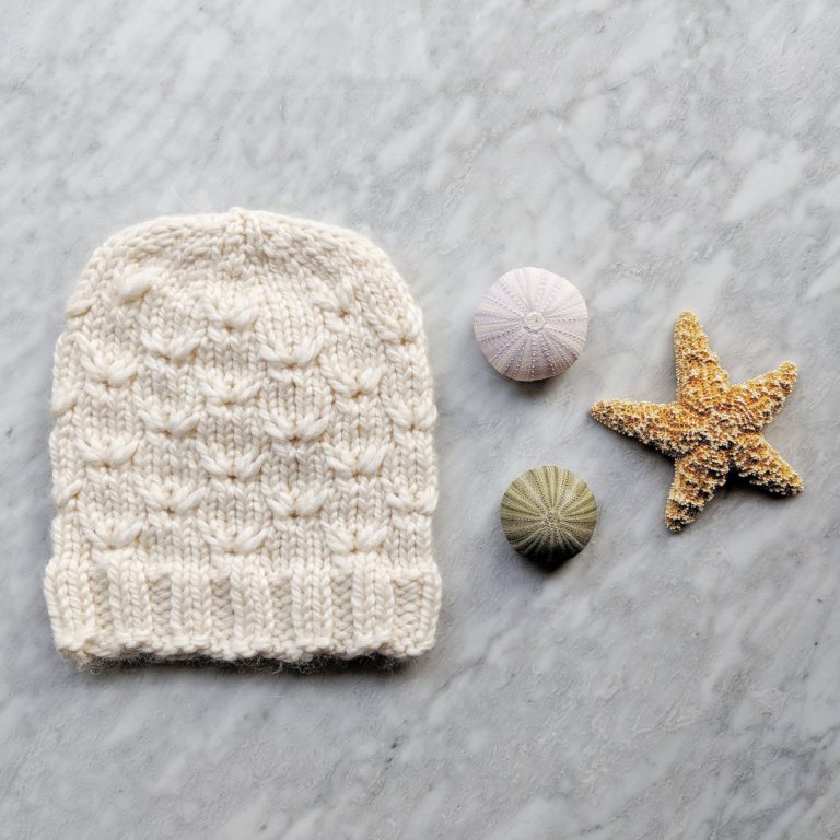 A white hat, knit with thick yarn in a cable design, lays on a grey marble countertop next to two sea urchins shells and a starfish.