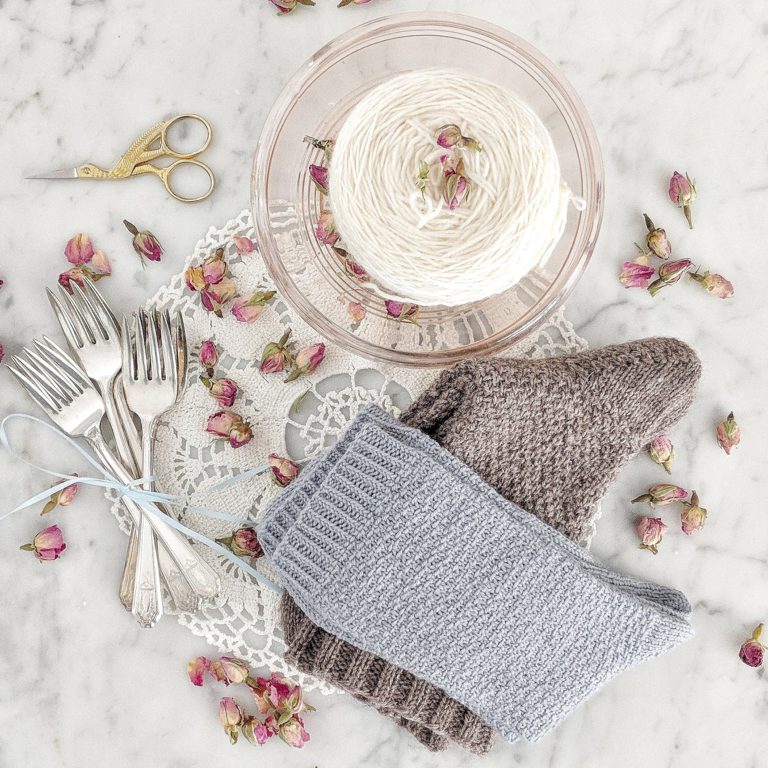 A pair of hand knit socks - one blue, one grey - are folded on a marble countertop atop a lace doily. A set of silver spoons tied with ribbon and a glass bowl with white yarn sit next to them. Dried rosebuds are scattered over the counter.