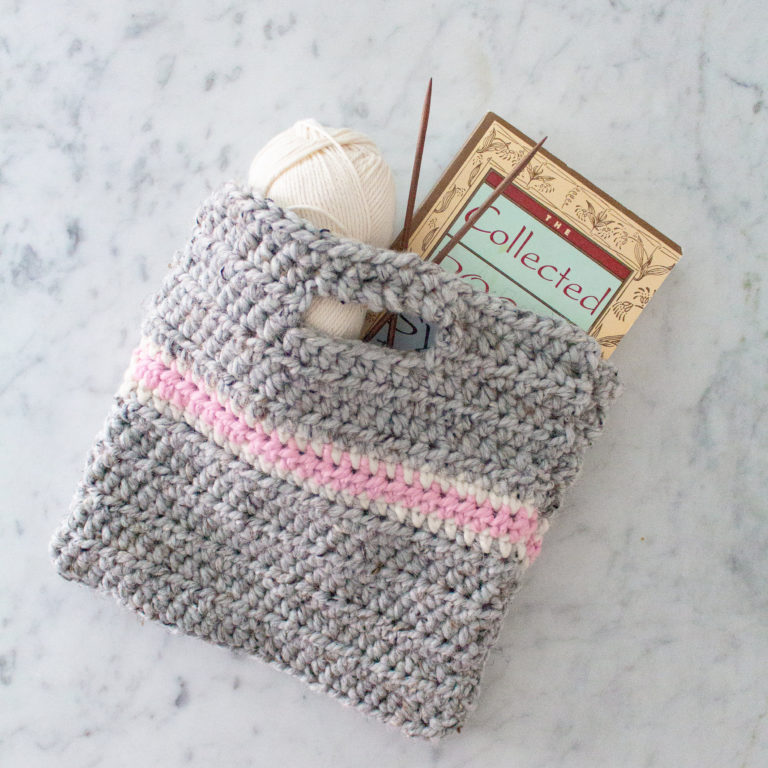 A small grey and pink crochet bag holds a white ball of yarn, two knitting needles, and a book. It rests on a marble countertop.