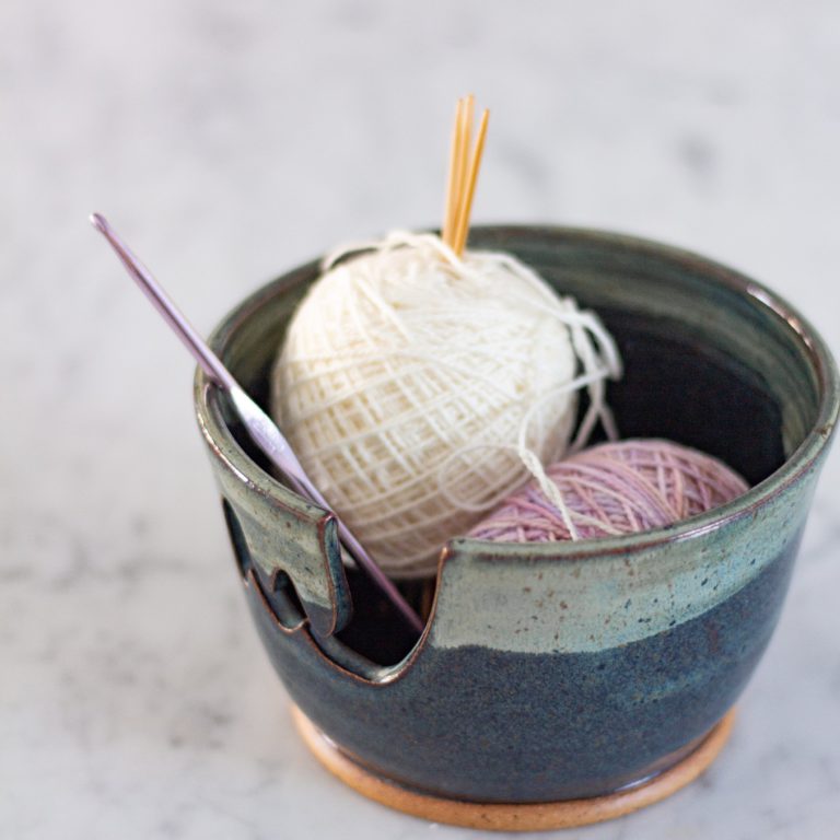 A handmade blue and green yarn bowl holds one white ball and one pink ball of yarn, as well as a purple crochet hook and three double-pointed knitting needles.