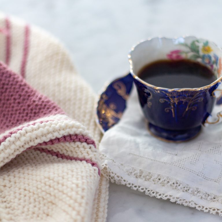 A blue china teacup with coffee in it sits on a lace doily. White and pink fabric, knit in garter stitch, lays next to it.