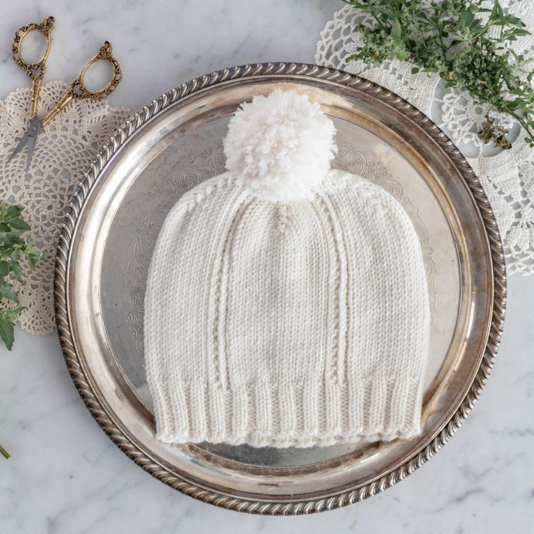 A white hand knit hat with a pom-pom lays on a silver platter. Lacy doilies and sprigs of wildflowers surround the platter.