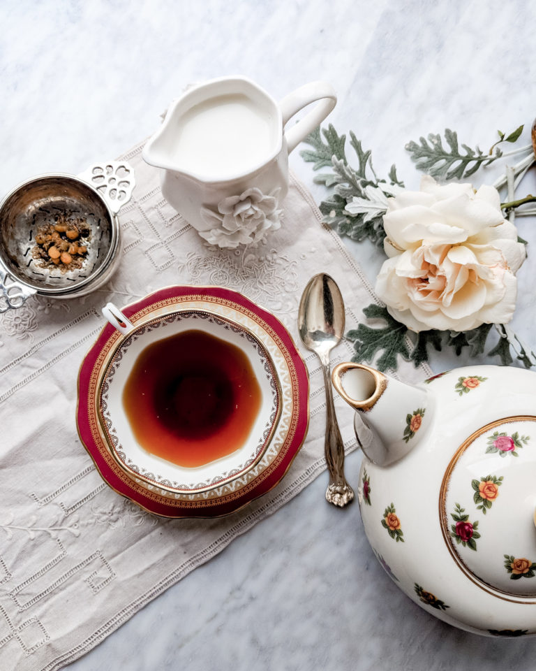 A red and white china teacup, partially filled with tea, sits on a lace placemat. A flowery teapot, a silver tea strainer, and a creamer with milk surround the teacup. A white-pink rose can be seen in the background.