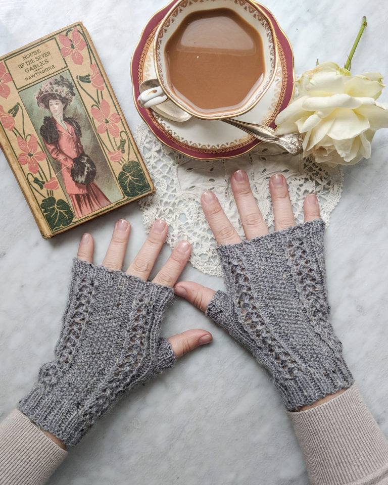 A white woman's hands, wearing a pair of grey hand knit fingerless mitts, are placed on a marble countertop. Above her fingers is a china teacup, a white rose, and a vintage book.