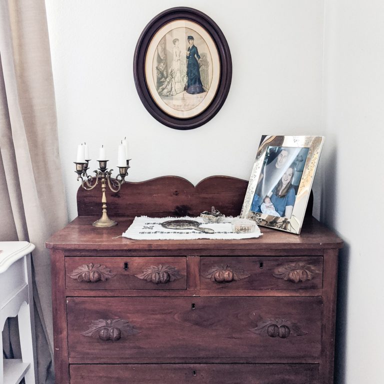 A vintage dresser, made of dark wood with decorative handles, sits under a round framed painting. A brass candelabra, a silver mirror, and a framed photograph rest atop the dresser.