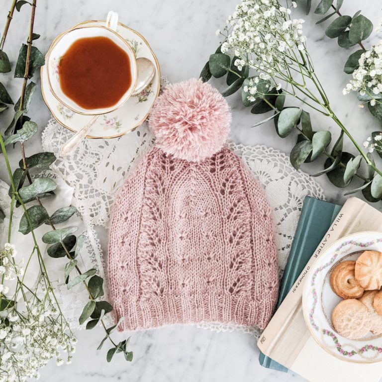 A pink hat, knit in a lace design with a pompom, rests atop lace doiliers. A china teacup sits to its left, and a stack of books with a plate of cookies on top is to its right. Springs of wildflowers surround the hat.