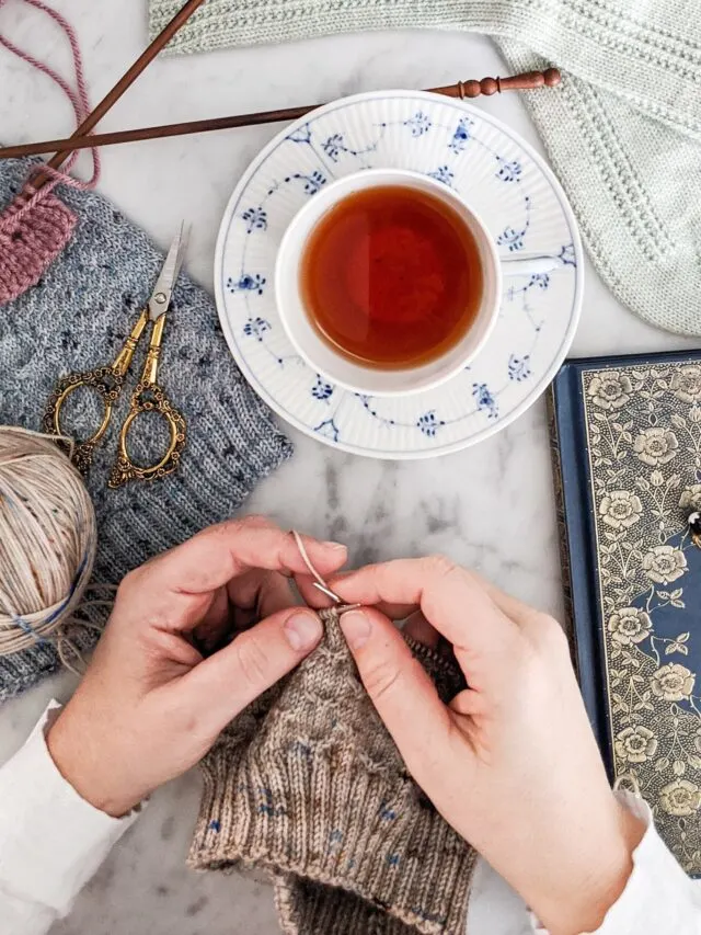 A pair of white hands works on a hat knit with light brown yarn speckled with pink and blue. They are surrounded by other knit items, a teacup filled with tea, a journal with a gilded cover, and ornate scissors. This beginner's guide to knitting will help you get started on your knitting journey.