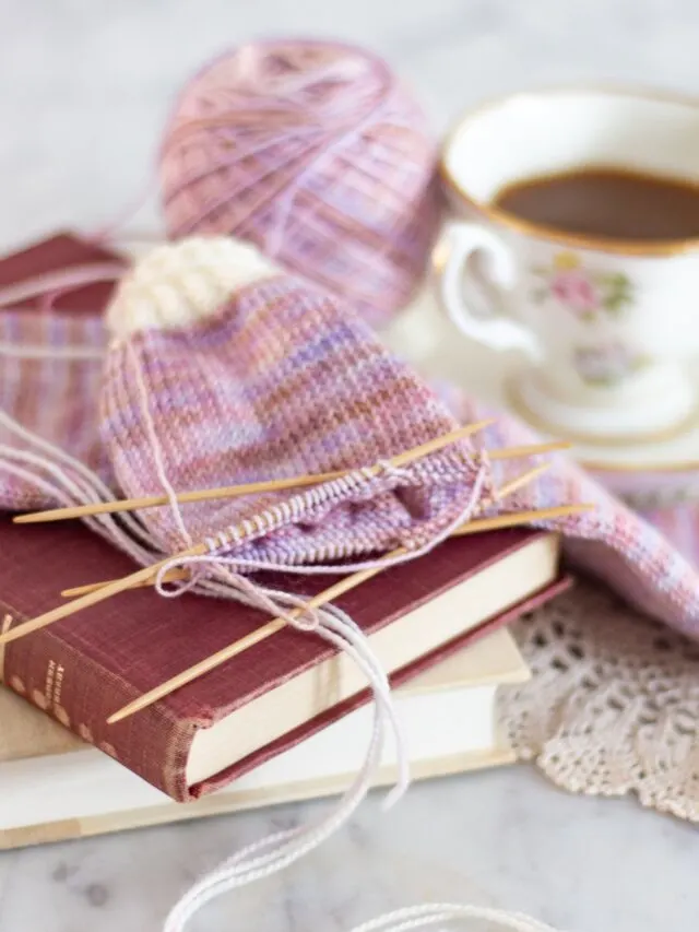 A partially-finished pair of variegated pink and white socks sit on top of a small stack of books. A teacup is slightly blurred in the background. Yarn substitution helped me learn how to use different yarn for socks, sweaters, hats, and more.