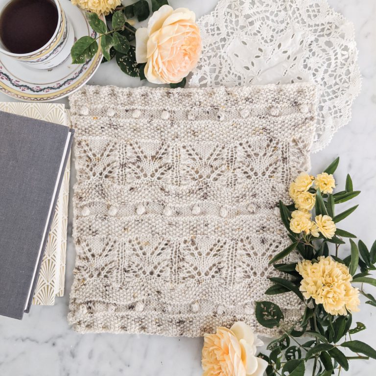 A light speckled cowl, knit with lace and bobbles, lays atop a marble countertop. Yellow roses, a stack of books, and a china teacup can be seen in the background.