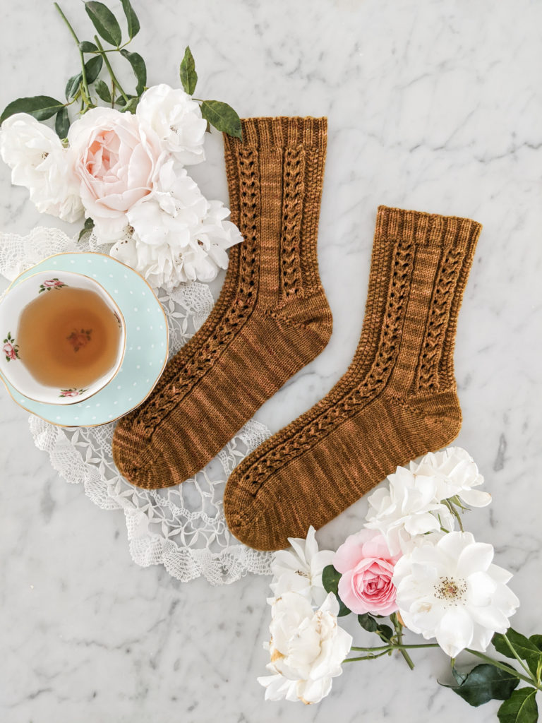 A pair of brown socks, knit with eyelet and seed stitch rows, sit on a marble countertop. White roses and a teal teacup sit next to the socks.