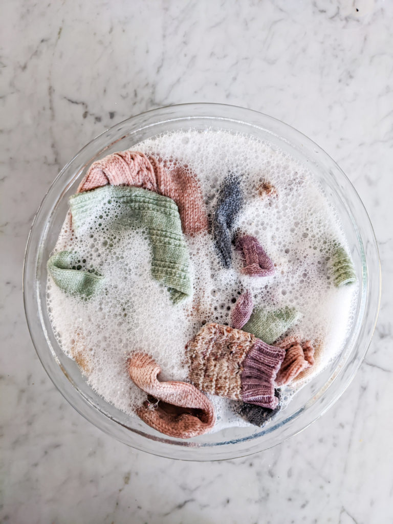 A glass bowl, filled with water and soap, holds multiple pairs of hand knit socks, which peek up through the bubbles.