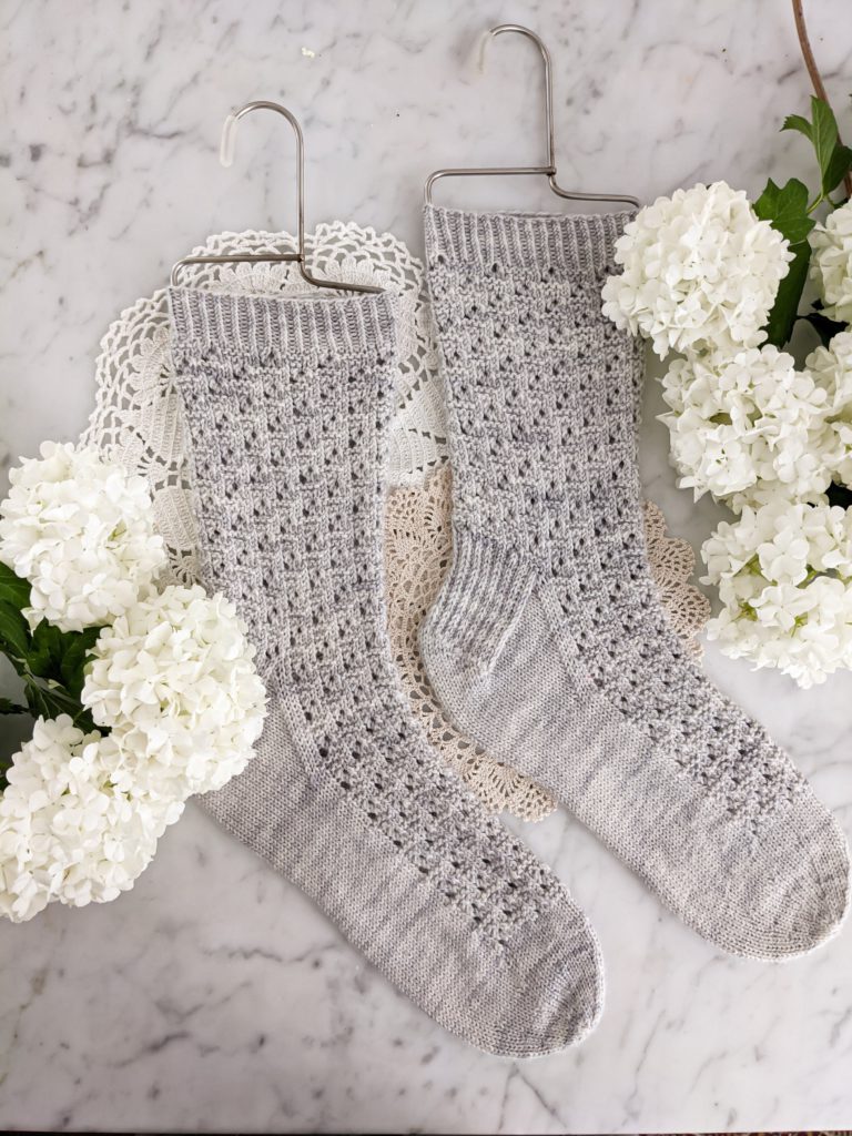 A pair of light blue-gray, textured socks is laid flat on a white marble countertop with some white hydrangea next to them. These socks are the Socktober 2020 Socks, a free sock knitting pattern.