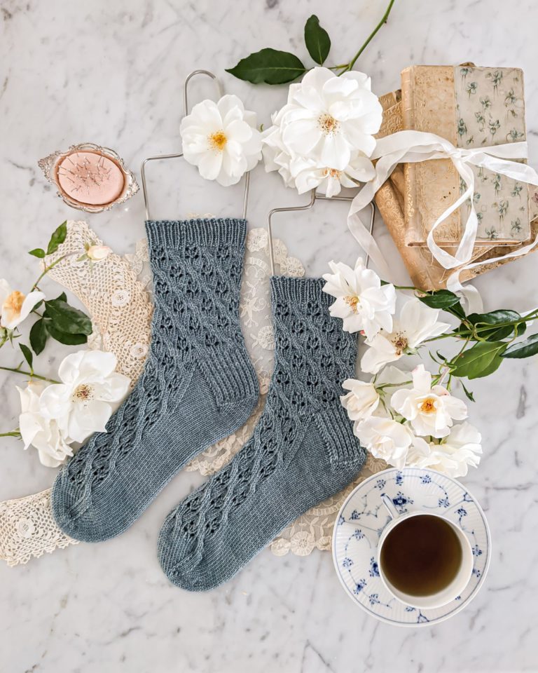 A pair of socks, knit with blue-grey yarn and a lace design, lay atop a lace doily. White flowers, a pink pincushion, old books, and a china teacup surround the socks.