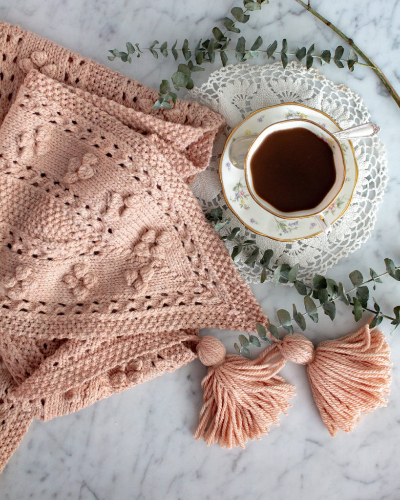 A pink scarf with pointed ends, lots of bobbles, and tassels is folded up on a white marble countertop. Next to it is a cup full of tea on top of a doily.