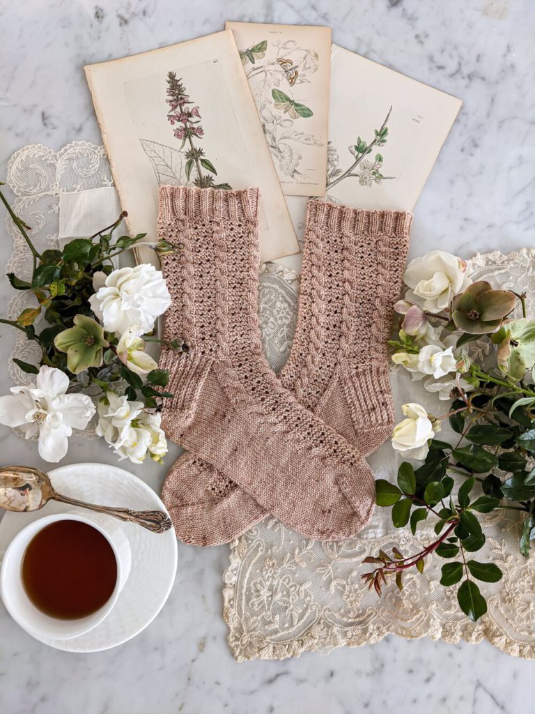 The Amicus Socks - A pair of pink socks with vertical cables and eyelets is laid flat on top of a cream lace background and some antique botanical prints. They are surrounded by white roses and white snapdragons, along with a cup of tea.