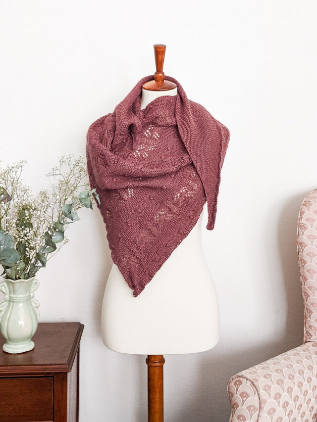 The Berry Scone Shawl, an asymmetrical triangle with stripes of lace, bobbles, and seed stitch, is draped around a white dressmaker's form. It's knit up in a desaturated burgundy color that's reminiscent of baked berries.