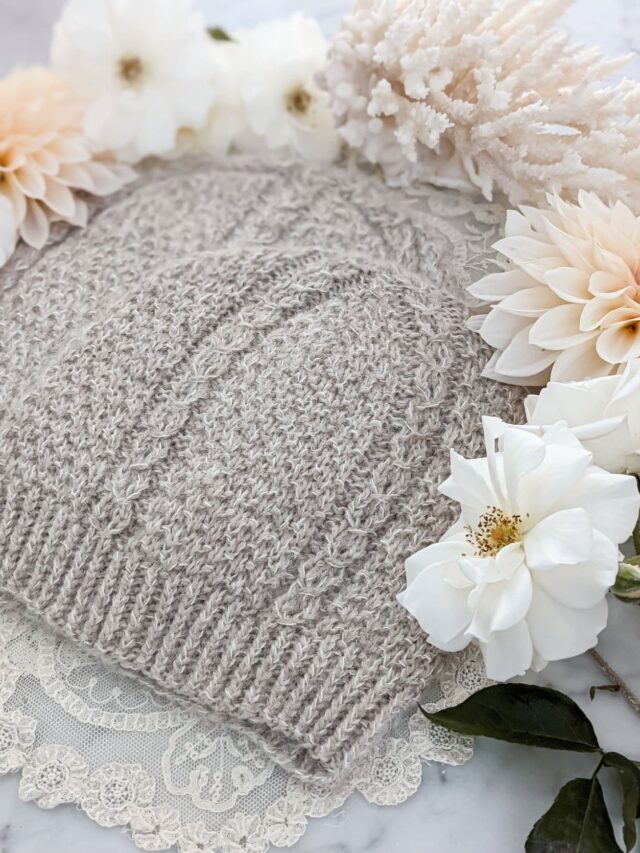 Two matching hats knit in fluffy tan yarn are laid flat on top of each other in a staggered fashion. They have seed stitch and columns of coin lace. They're surrounded by peach dahlias, white roses, and spiky white coral.