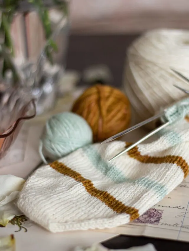 A white, caramel, and mint striped sock toe is laid flat with small balls of mint and caramel yarn behind it. The sock is knit on double-pointed needles.