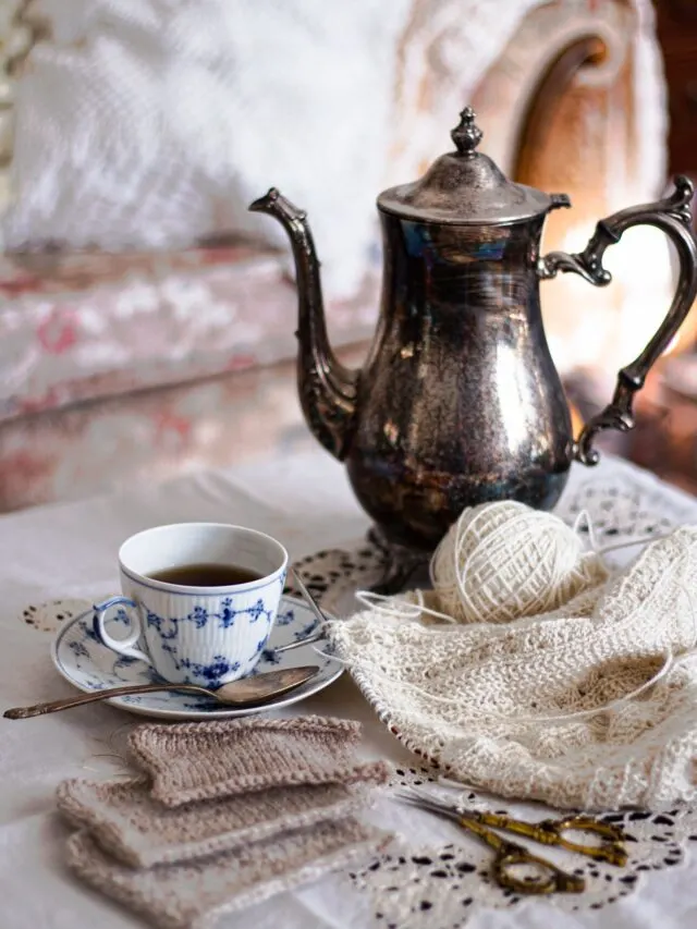 In the foreground are ornate brass scissors and three tan swatches of knit fabric. The middle distance in focus features a pile of creamy white knitting, a blue and white teacup and saucer, and a silver coffee pot. Blurred in the background is a pale floral sofa.