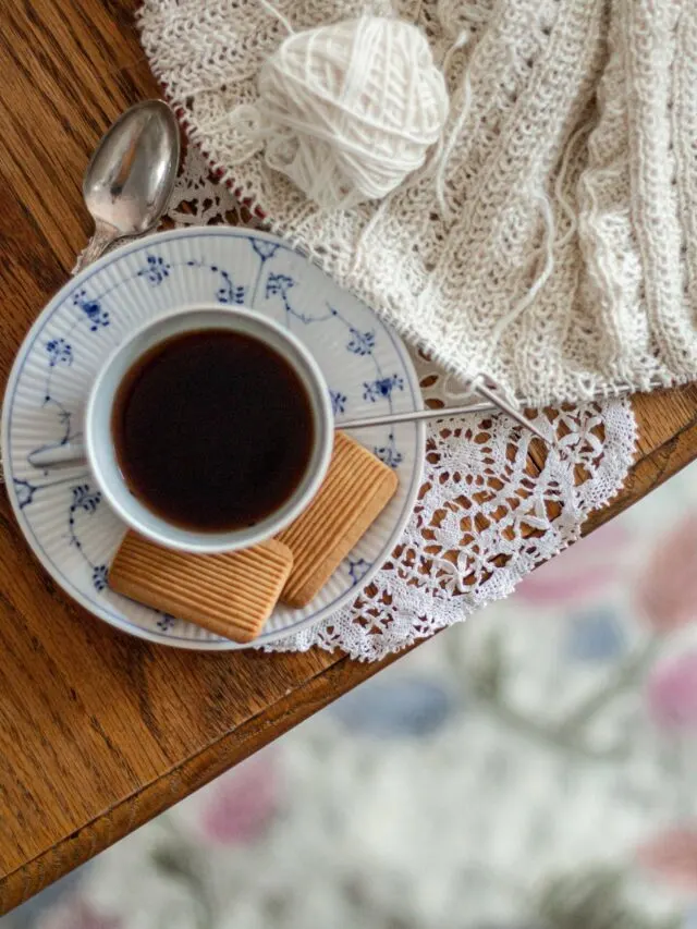 A top-down shot of a blue and white teacup and saucer sitting on the edge of an oak table with a white doily and some white knitting on steel needles. Knitting helps me overcome perfectionism by giving me a safe place to make mistakes.