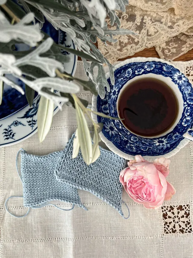 Two blue rectangle swatches are viewed top down through blurred plant leaves in the foreground. One swatch has even stitches, while one has visible rowing out. They are on a white linen placemat next to a blue and white teacup.