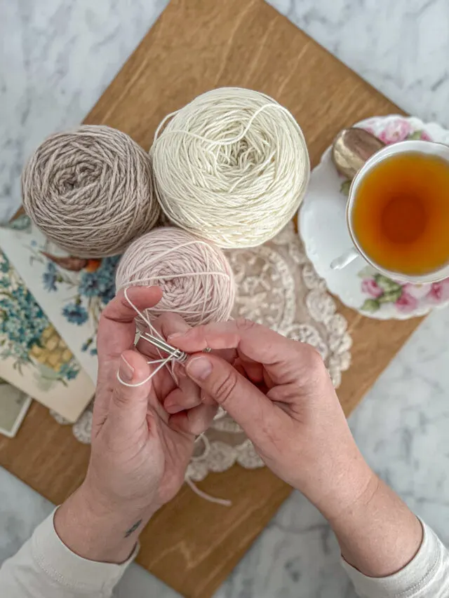 A top-down view of two small, plump, white hands casting on yarn over two needles. Also in the photo is a wooden board, some balls of pastel yarn, and a pink and white teacup.