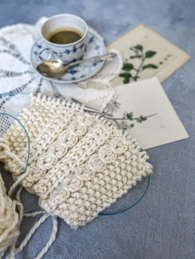 A close-up image of a cream-colored swatch of knit fabric. Behind it, blurred in the background, are a teacup, a doily, and some antique botanical prints.