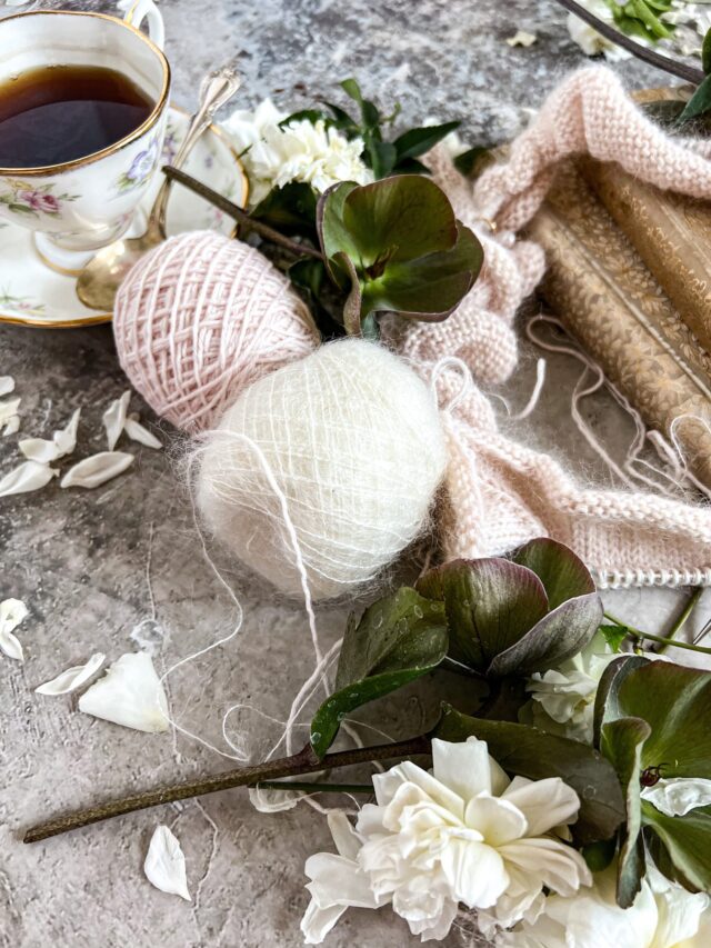 A close-up on two balls of yarn, one pink and one white, surrounded by flowers, a teacup, and some knitting