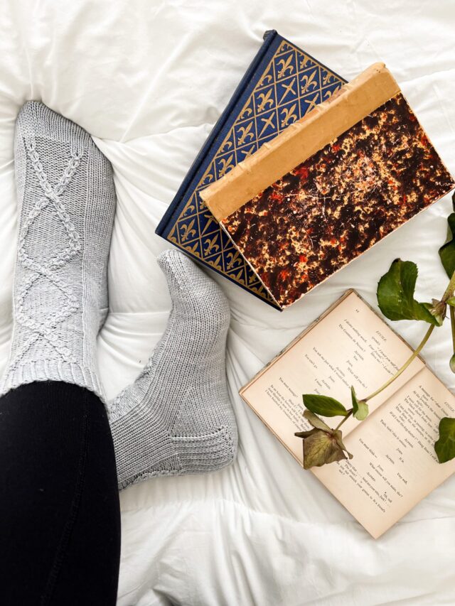 The Deimatic Socks, a pair of light blue, cabled socks on feet that are crossed at the ankle. Next to them are some antique books.