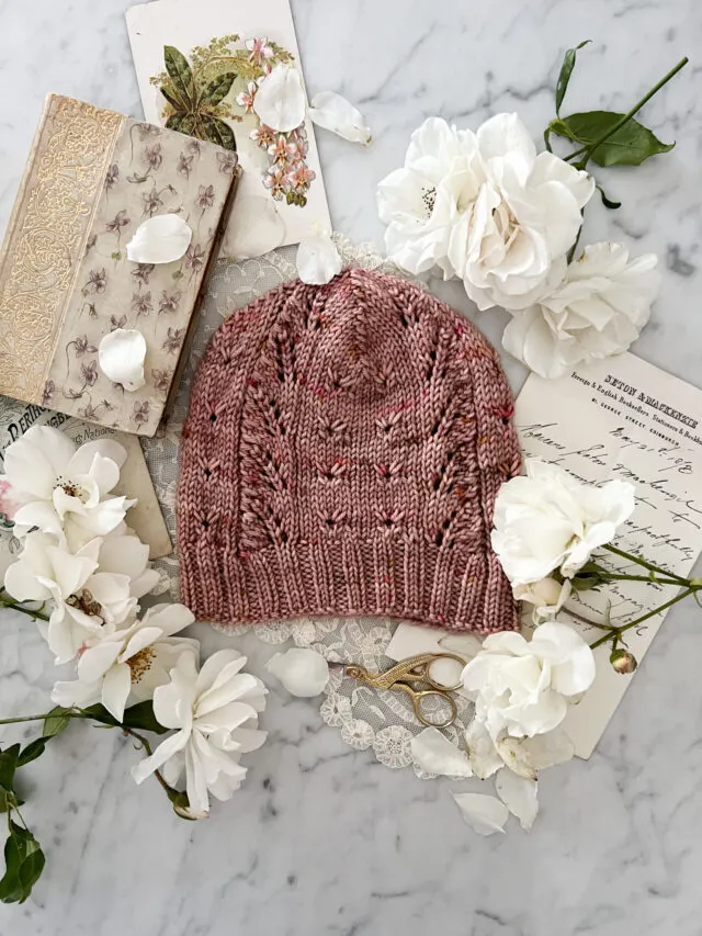 A pink hat with lace panels and eyelets is laid flat on a white marble countertop, surrounded by white roses, antique paper ephemera, and an old book with a gilded cover.