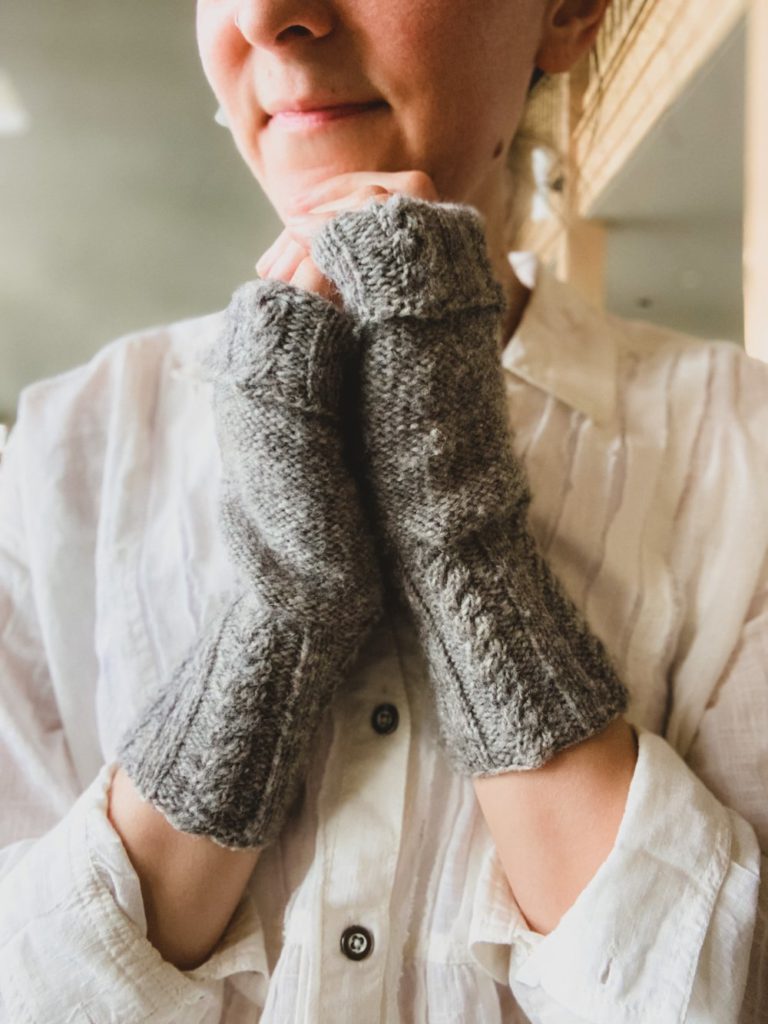 A pair of gray fingerless mitts with cabled cuffs on a woman's hands clasped together in front of her chin.