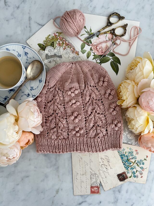 A Top-down view of the Laurel Hat, a pink hand knit hat with bobbles and lacy leaves, which is laid flat on a white marble countertop. It's surrounded by roses, a teacup full of espresso, and antique paper ephemera.