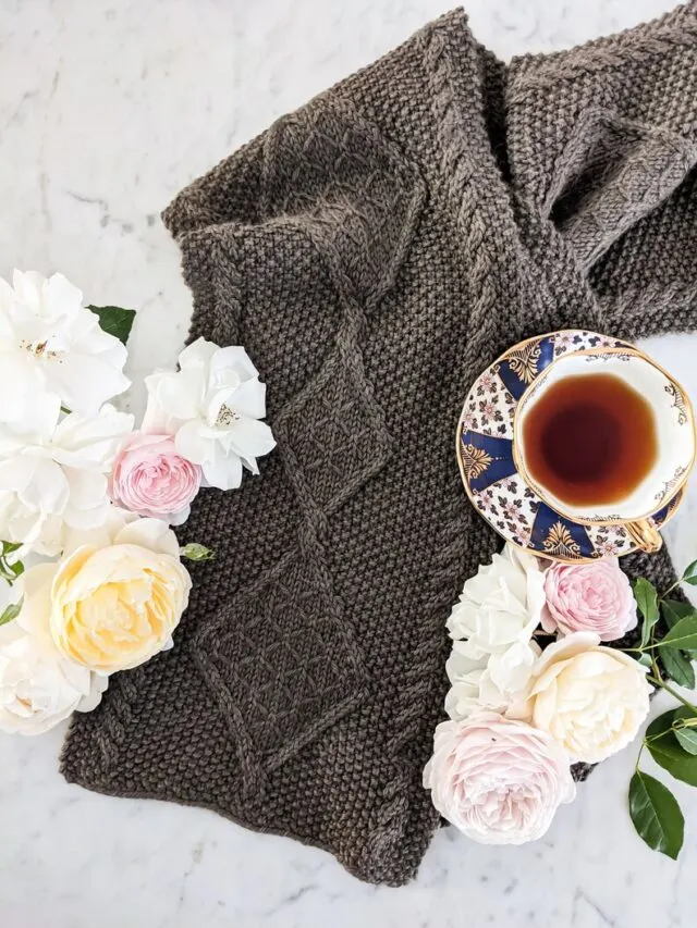 A brown cabled scarf is laid flat on a white marble countertop. It is surrounded by pink, white, and yellow roses and a teacup full of Earl Grey.