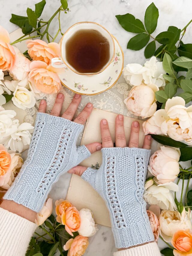 A woman's small, plump hands are splayed slightly on a white countertop. They're wearing a pair of light blue fingerless mitts with panels of lace and seed stitch down the back of the hands. Around them are lots of peach and cream roses. A teacup full of tea sits at the top of the photo.