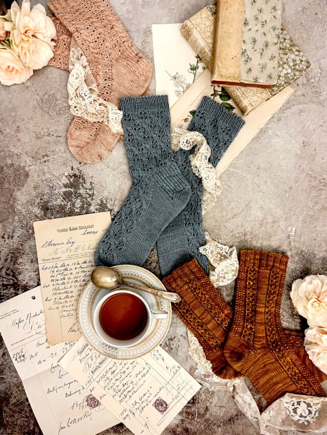 Three pairs of knit socks (pink, blue, and caramel) are laid out on a flat surface in a diagonal from top left to bottom right. They're surrounded by white roses, antique books, a teacup full of chamomile, and antique paper ephemera.