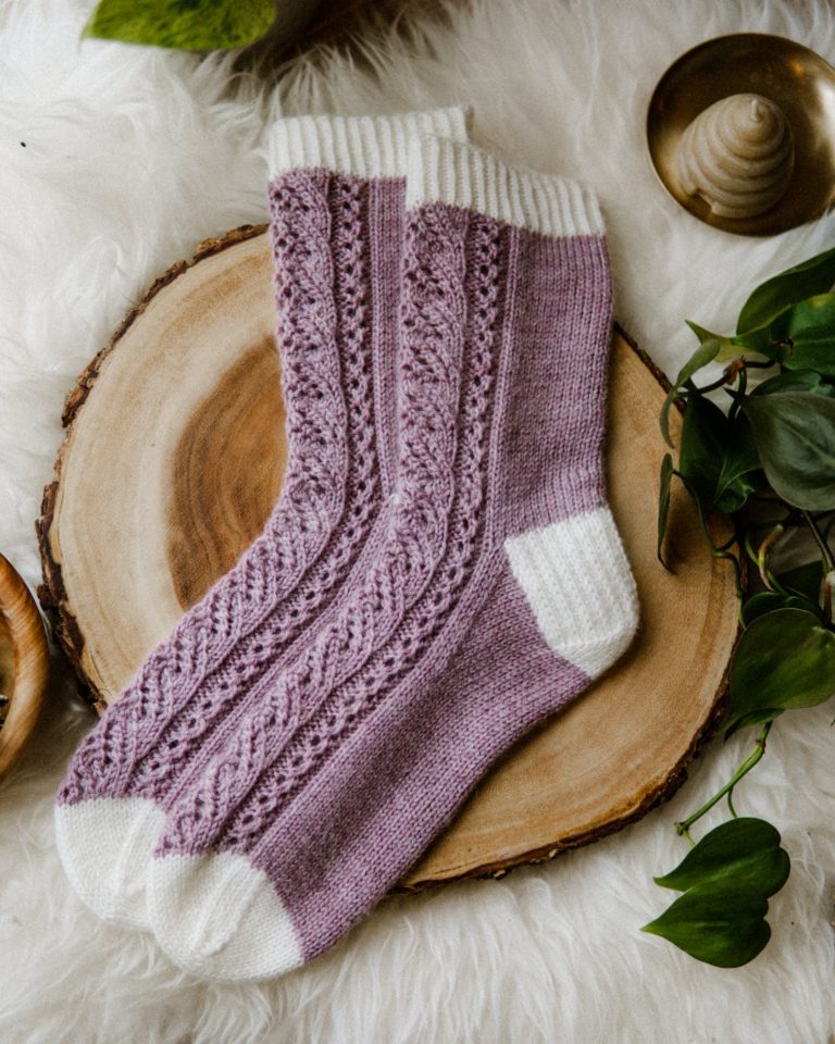 A pair of purple lacy socks with white cuffs, heels, and toes is laid out on a thin sliver cut from a tree trunk. The toes are pointing to the left.