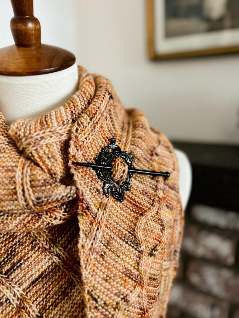 A close-up on a shawl pin made of a brass circle of art nouveau-style calla lilies and a wooden stick pin.