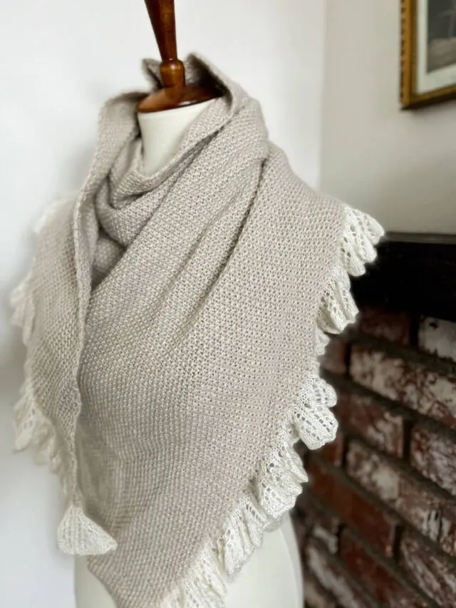 A side angle of the Grandmillennial Shawl draped around a dressmaker's form. It's a seed stitch shawl knit in a gray cashmere-merino blend from Purl Soho and features a soft ruffle in a white silk-mohair yarn.