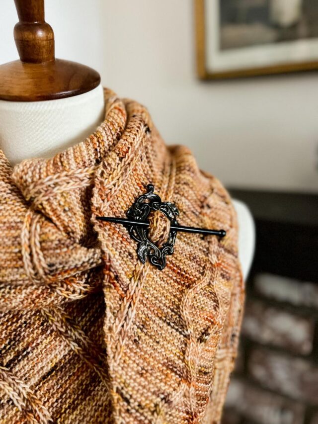 A close-up on a shawl pin made of a brass circle of art nouveau-style calla lilies and a wooden stick pin.