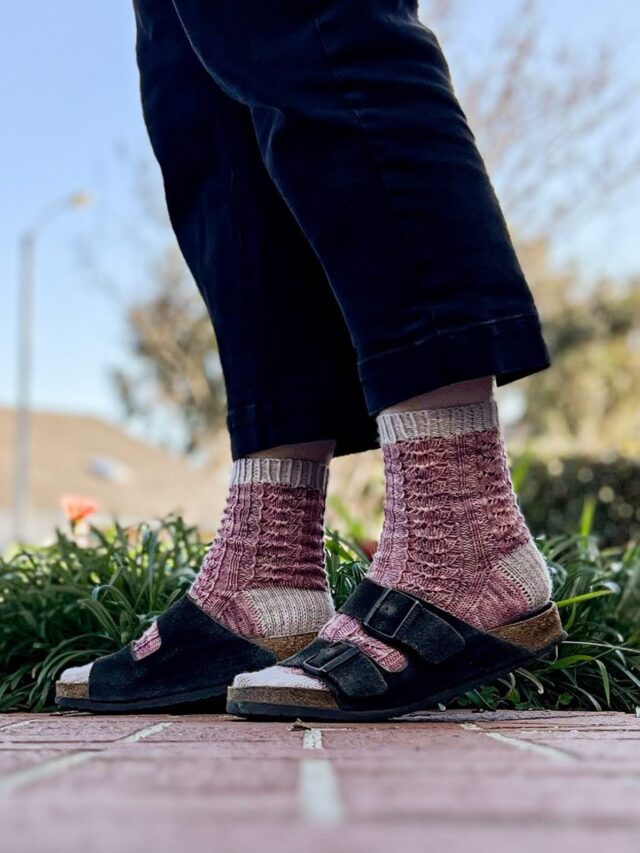 A pair of pinky purple lace socks with gray cuffs, heels and toes are worn by a woman with Birkenstock sandals over them. The photo focuses on her feet, which are mid-step. The photo is taken outside on a sunny day.