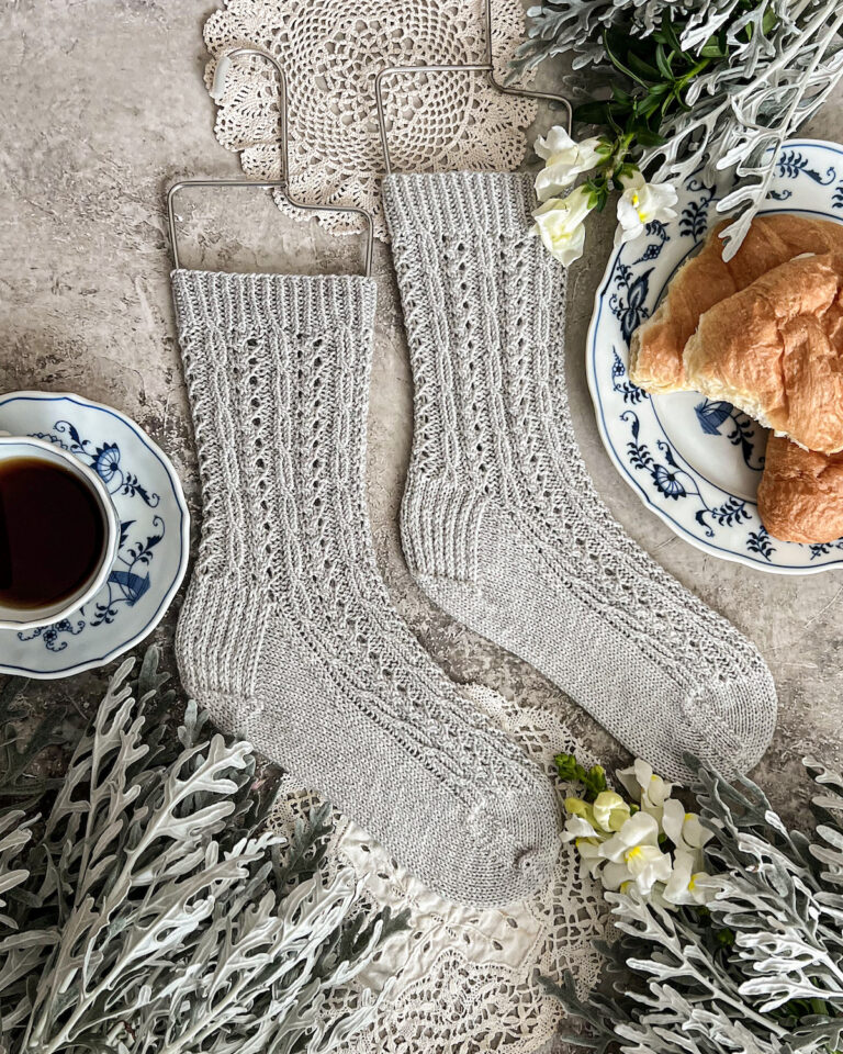 A flatlay photo of a pair of light gray handknit socks. The socks are laid flat on a gray surface with the toes pointing to the right. They're covered in tiny cables and eyelets, and surrounded by dusty miller, a blue and white teacup full of coffee, and a blue and white place with several croissants on it.