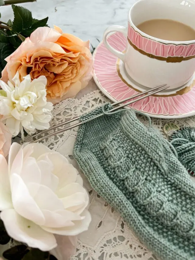 A close-up on the toe of an almost-finished sock. The toe stitches are arranged on two needles and ready to be grafted together. The sock is surrounded by a pink teacup full of milky tea and some peach and white roses.