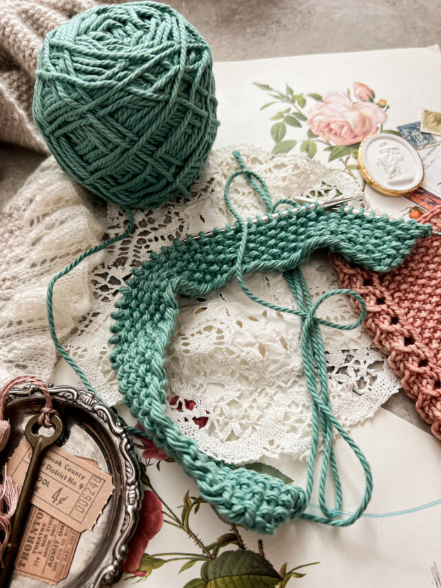 A turquoise dishcloth in progress fills the middle of the photo. It's about an inch tall and curves along a pair of circular needles. Cropped at the edges are a pink cowl and a tiny silver tray full of trinkets.