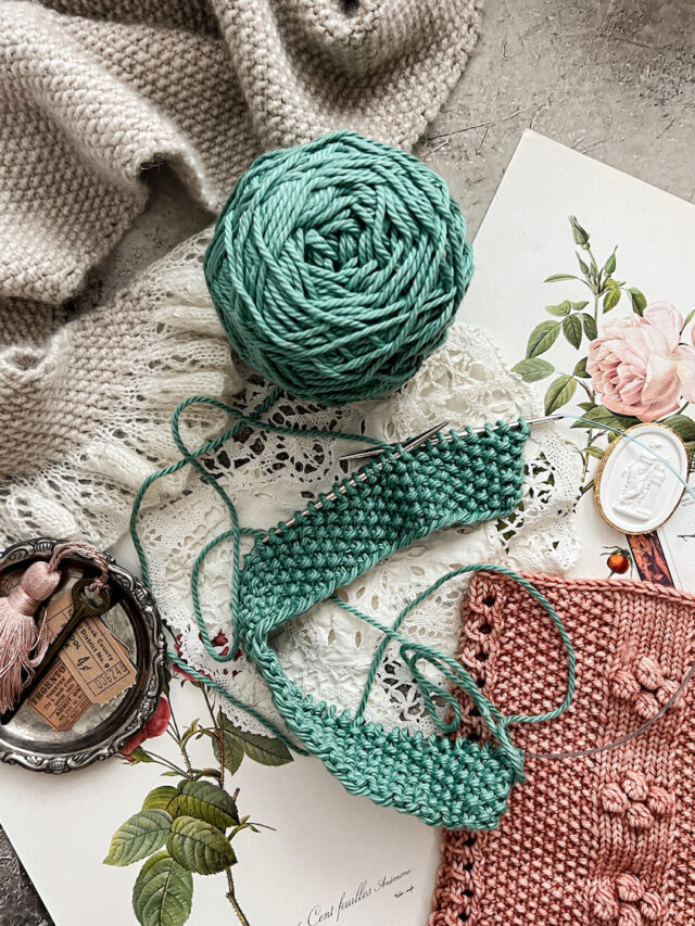 A horizontal photo showing three projects made with seed stitch: a gray shawl at top left, a pink cowl at bottom right, and a turquoise dishcloth in progress in the middle. they're surrounded by antique paper ephemera and trinkets.