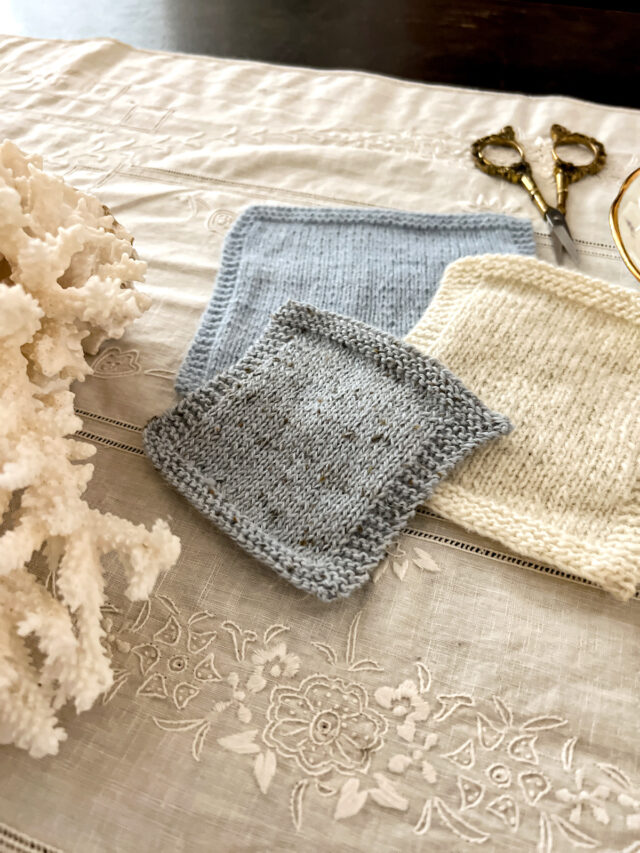 Three swatches of knit fabric in gray, white, and blue are laid out on an antique table cloth. A chunk of coral to the left is cut off by the edge of the photo frame.