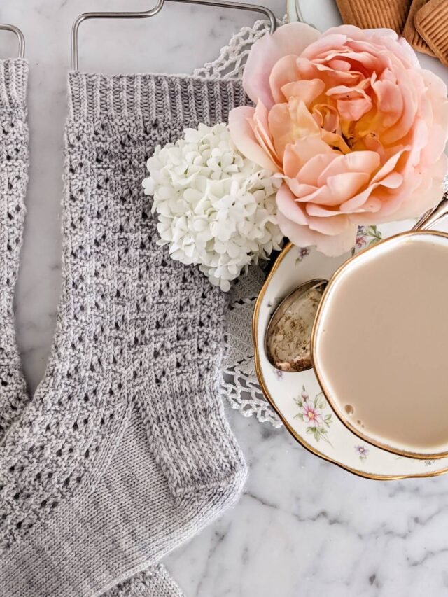 A close-up on the leg and heel of a light gray sock with eyelet texture. To the right is a peach rose, a white viburnum, and a teacup full of milky tea.