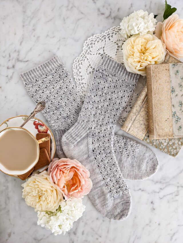 A flatlay photo of a pair of light gray knit socks surrounded by peach and yellow roses, antique books, and a teacup full of milky tea.