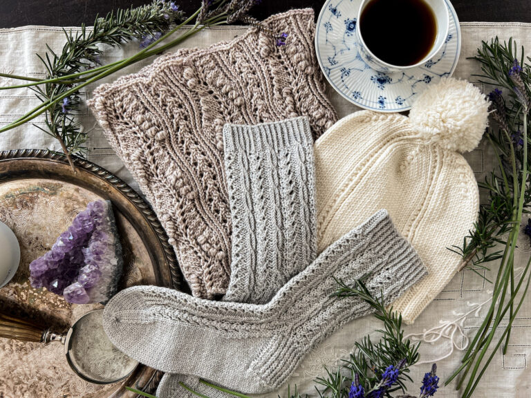 A hat, pair of socks, and cowl, all featuring twisted stitches as part of their stitch patterns, are laid on top of each other on an embroidered linen table cloth. They're surrounded by fresh greenery from the garden, a blue and white teacup full of black coffee, and a silver tray with some curios on it