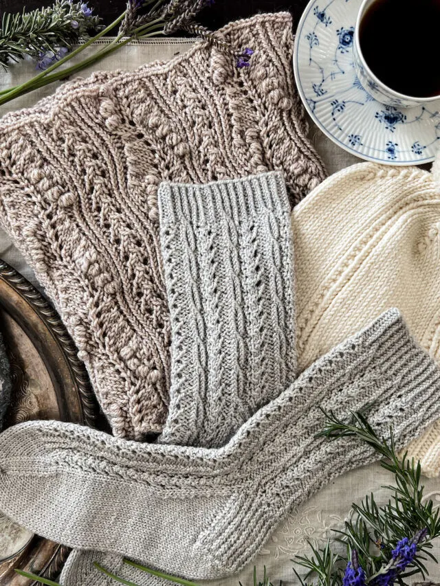 A hat, pair of socks, and cowl, all featuring twisted stitches as part of their stitch patterns, are laid on top of each other on an embroidered linen table cloth. They're surrounded by fresh greenery from the garden, a blue and white teacup full of black coffee, and a silver tray with some curios on it