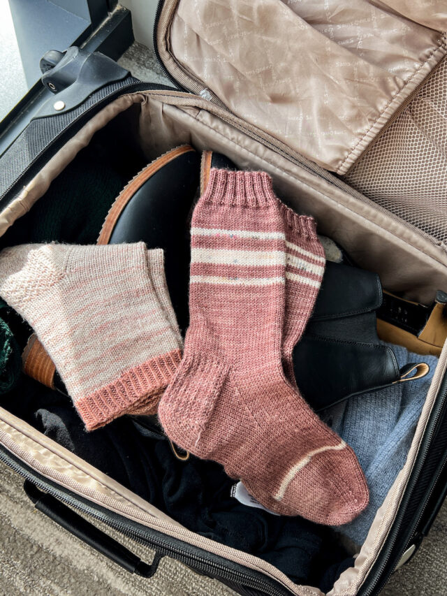 Two pairs of pink, hand-knit socks sitting in the top of an open suitcase.